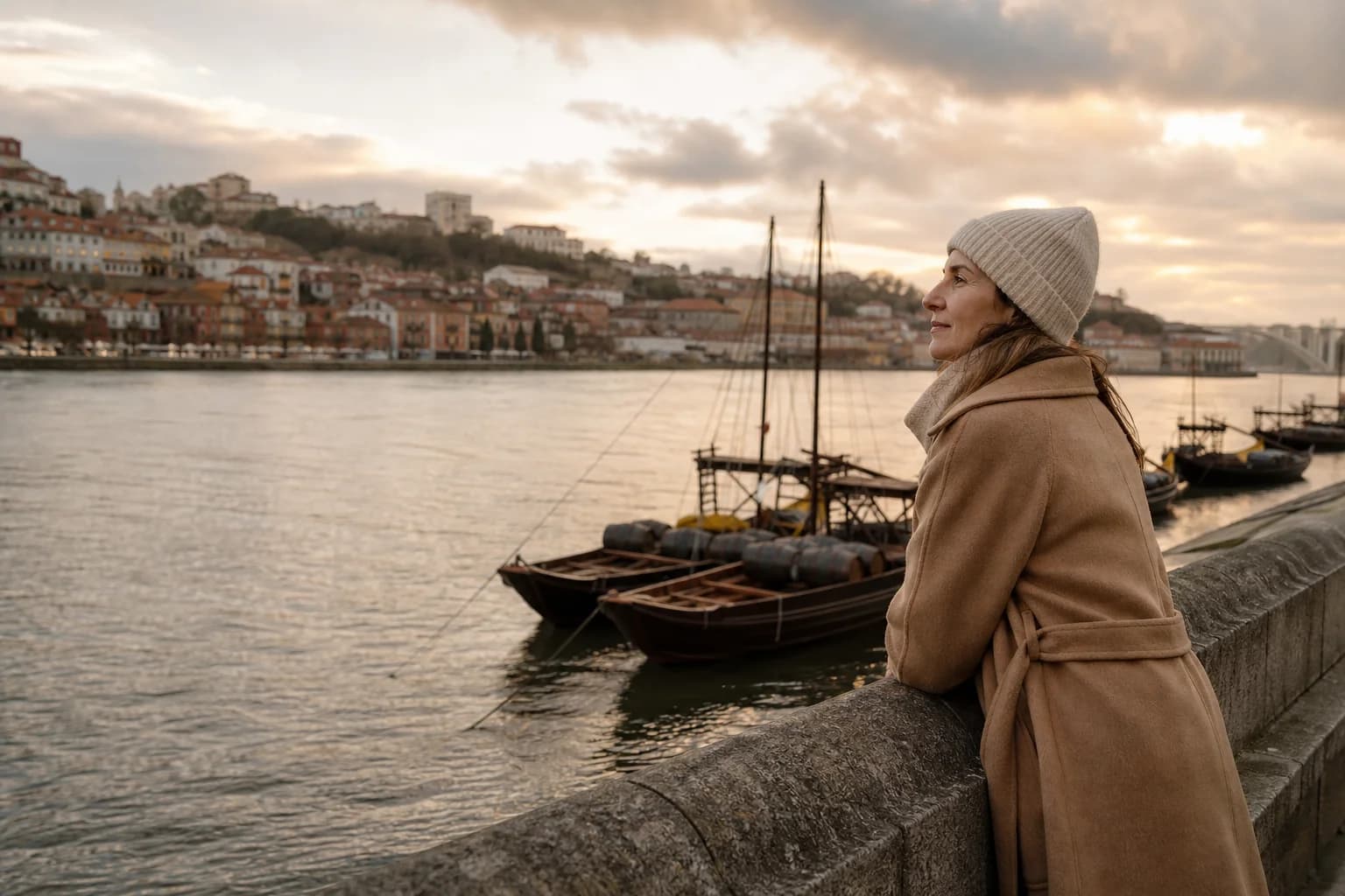 Mulher com casaco comprido cor de camelo encostada ao muro junto ao rio Douro, com barcos rabelos ao fundo